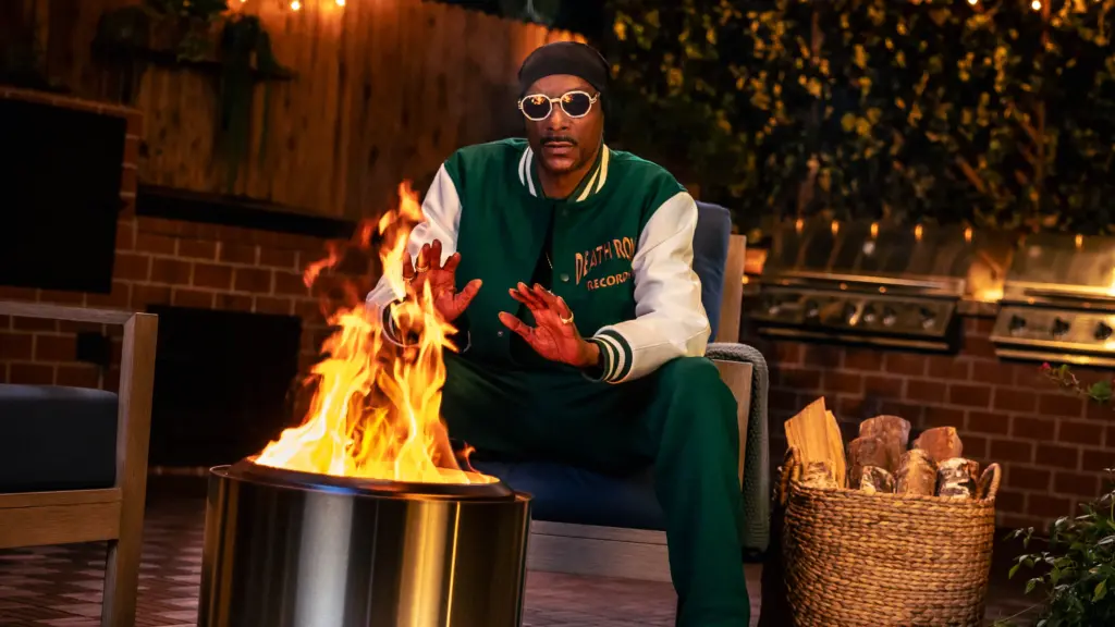 An image of the rapper Snoop Dogg wearing sunglasses warming his hands over an outdoor firepit.